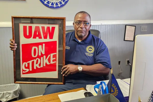 photo of ricky mcdowell sitting at his desk holding a framed sign that reads "UAW on Strike"