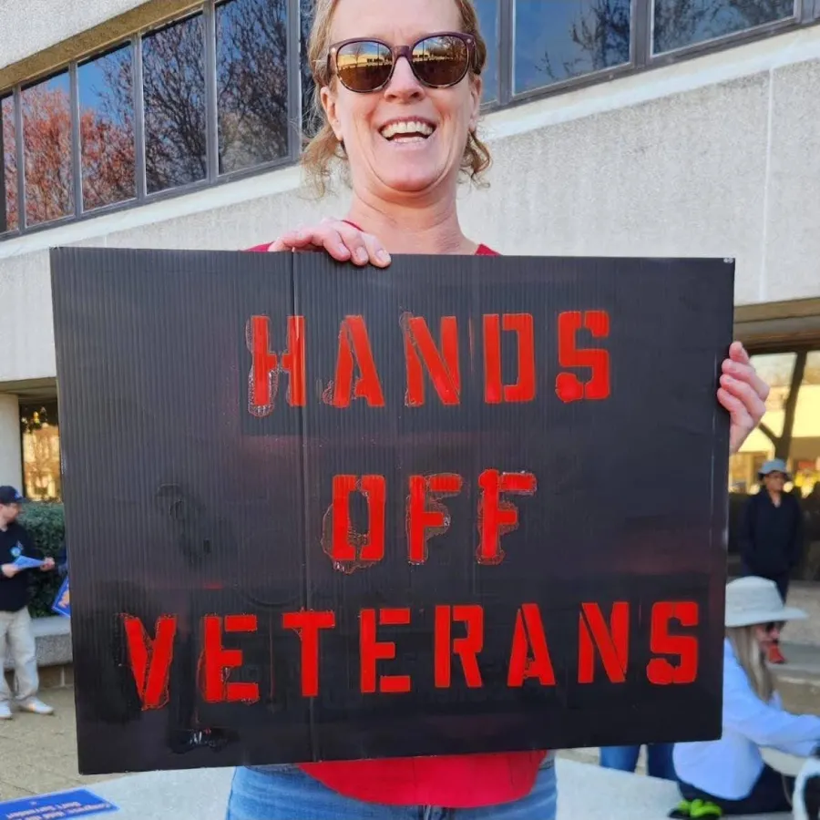 photo of libby manly holding a protest sign that reads "hands off veterans"