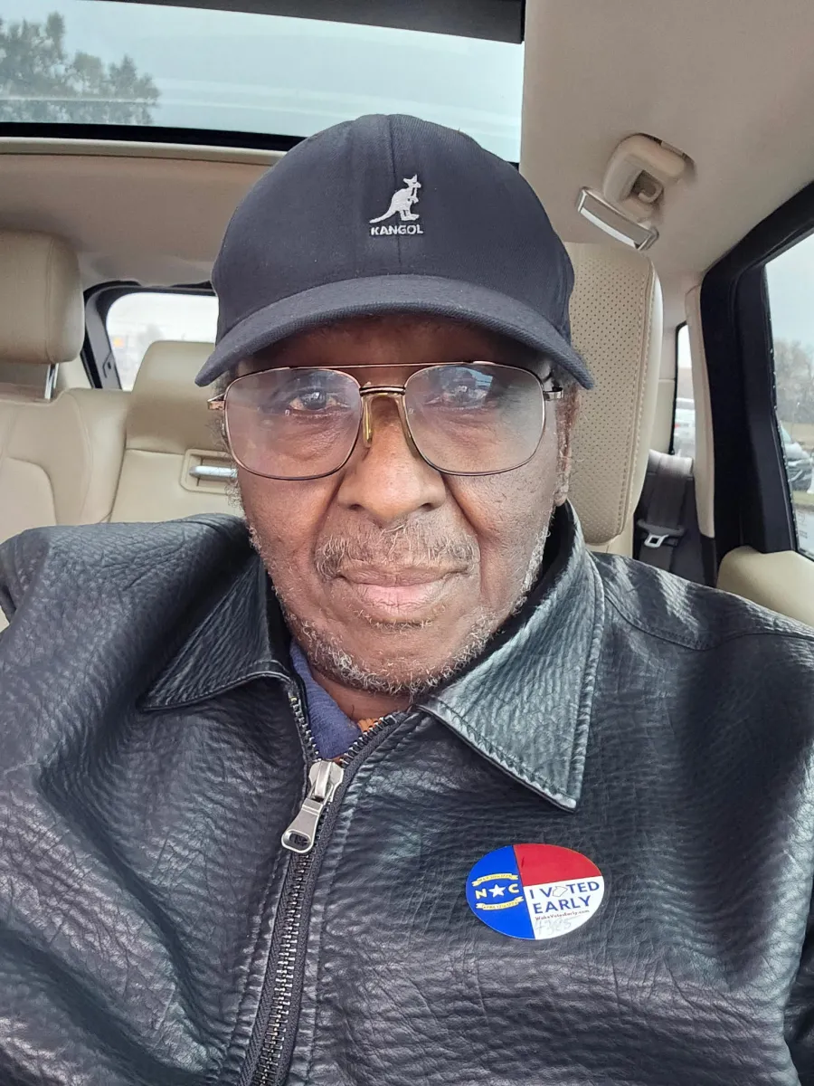 photo of Wayne Bostick in his car wearing glasses, a hat, and a leather jacket with a NC voter sticker on it