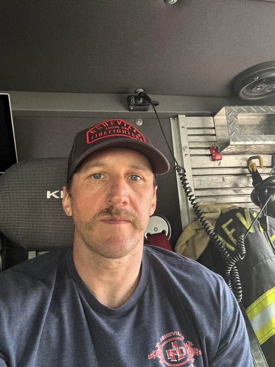 photo of Welcker Taylor wearing a hat and t-shirt that read Asheville Firefighters Local 332 and sitting in the cab of his fire truck