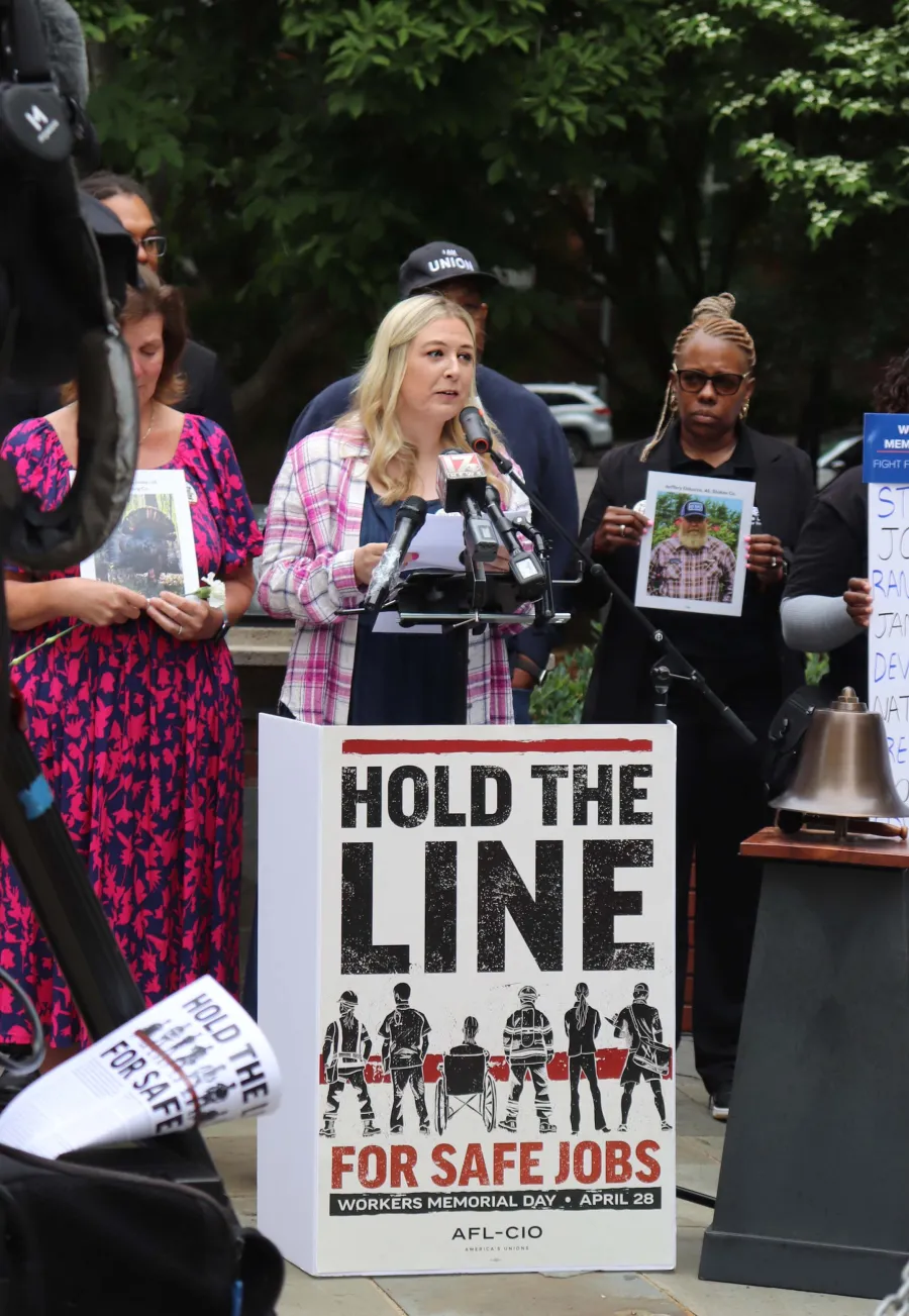 photo of Gianna Polito speaking at NC Workers Memorial Day service behind a sign that reads "hold the line for safe jobs"
